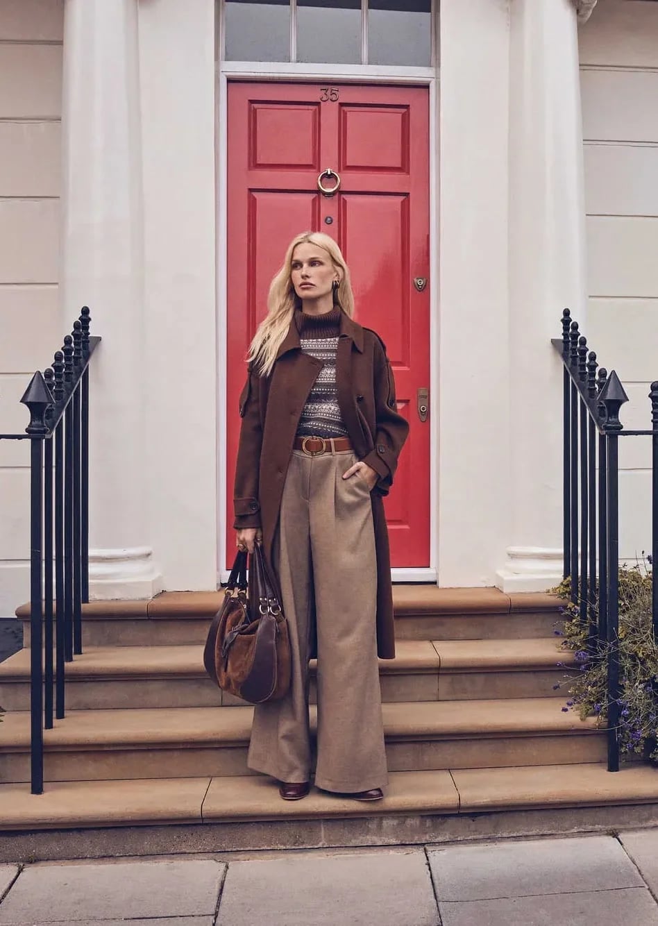 A woman in a long brown coat and wide beige trousers on the steps of a London house with a red door