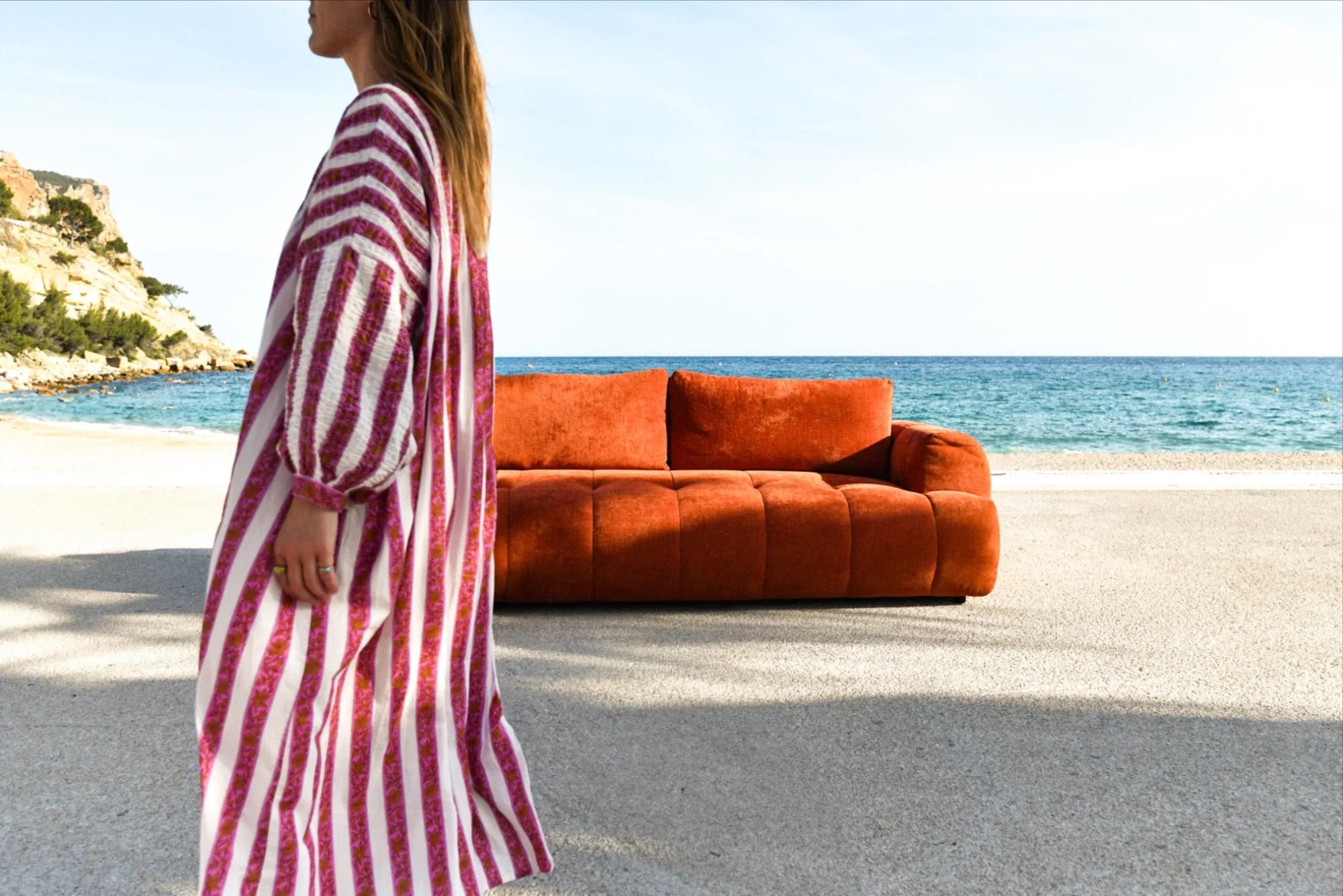 A woman in a pink striped dress walks along a beach, in front of an orange velvet Bobochic sofa facing the sea