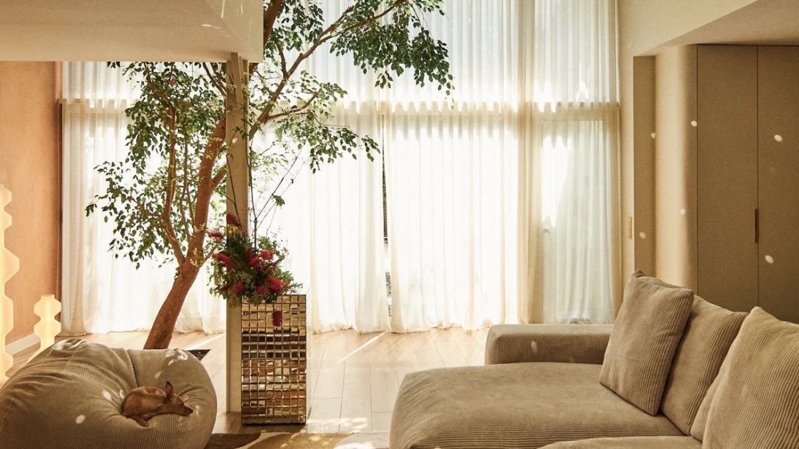 A bright living room furnished by Bobochic: a large beige corner sofa, a pouf, an indoor tree and long white curtains