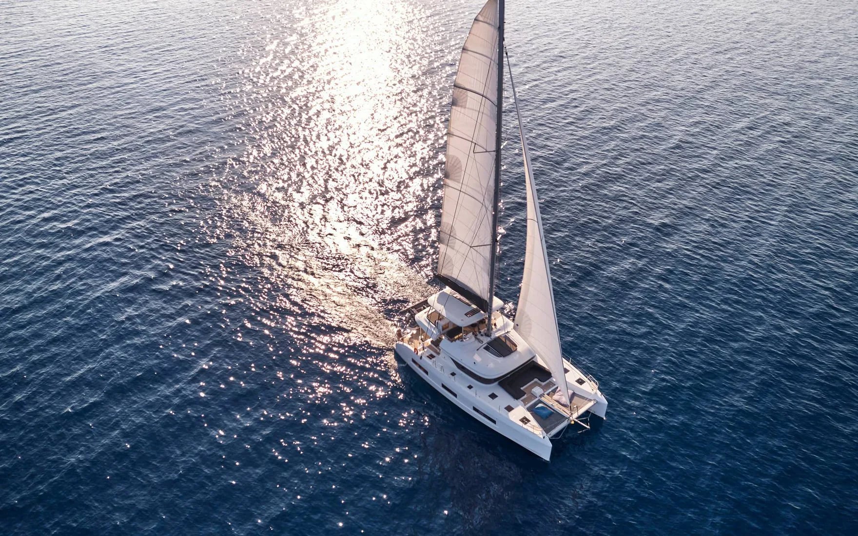 Un catamaran sous voiles sur une mer scintillante, vu du ciel à contre-jour