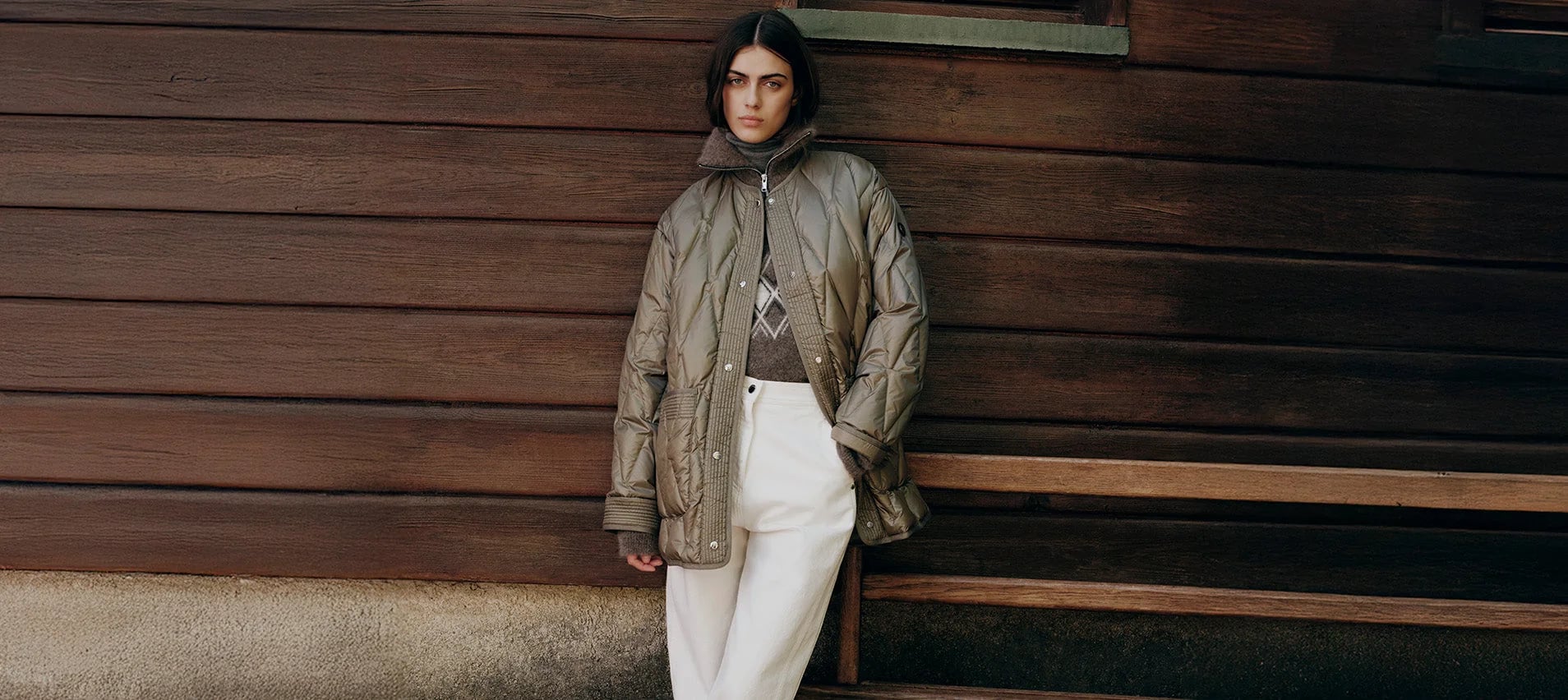 A woman in a Fusalp quilted jacket and white trousers, leaning against the wooden wall of a chalet
