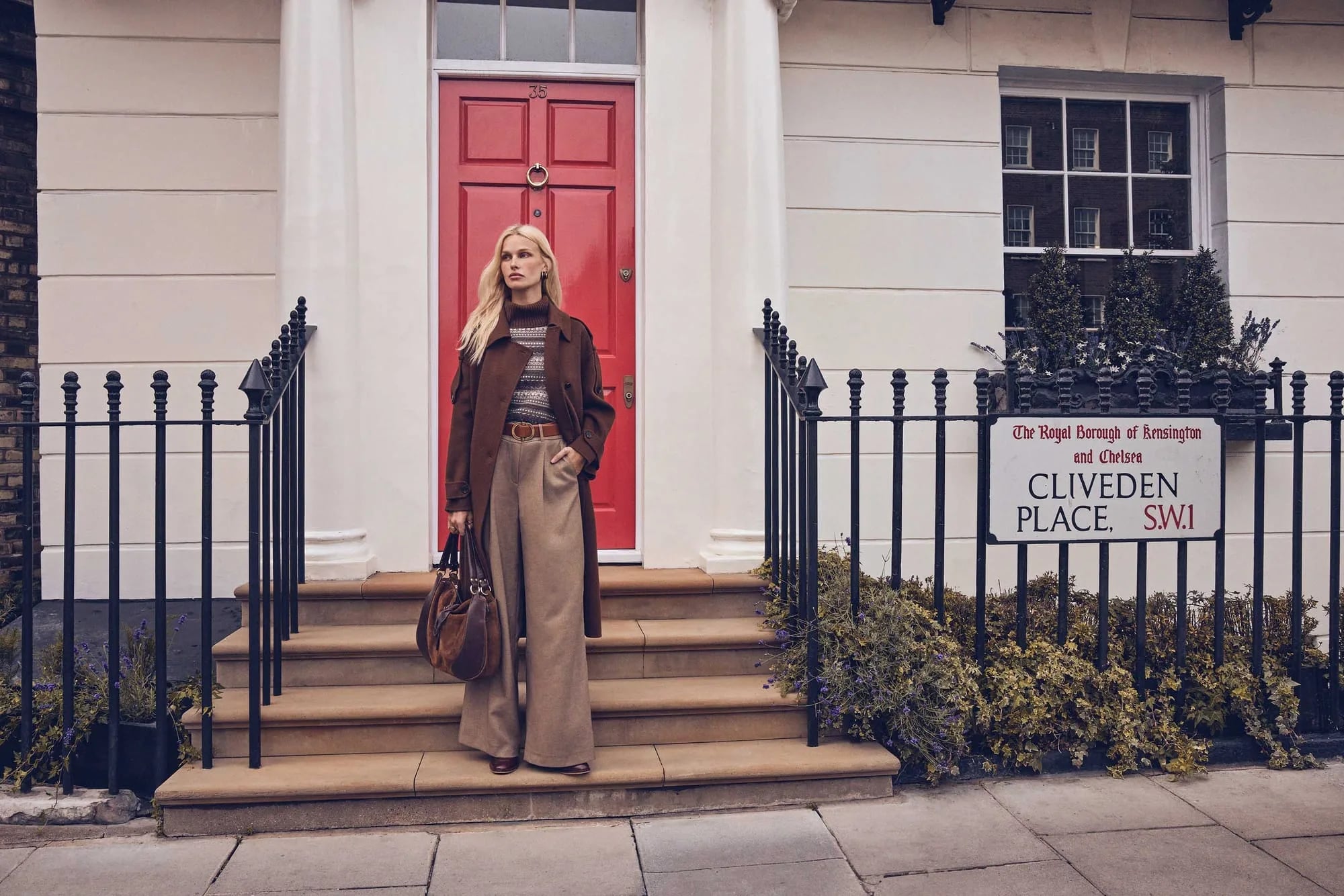 A woman in a long brown coat and wide beige trousers on the steps of a London house with a red door