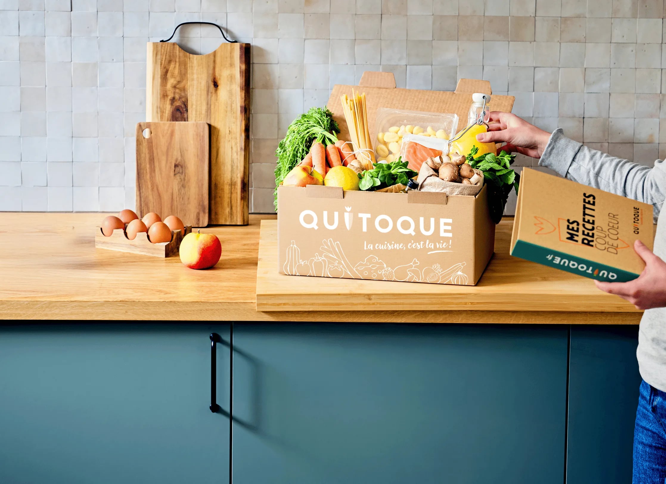 A Quitoque box filled with fresh vegetables on a kitchen worktop, next to a recipe booklet